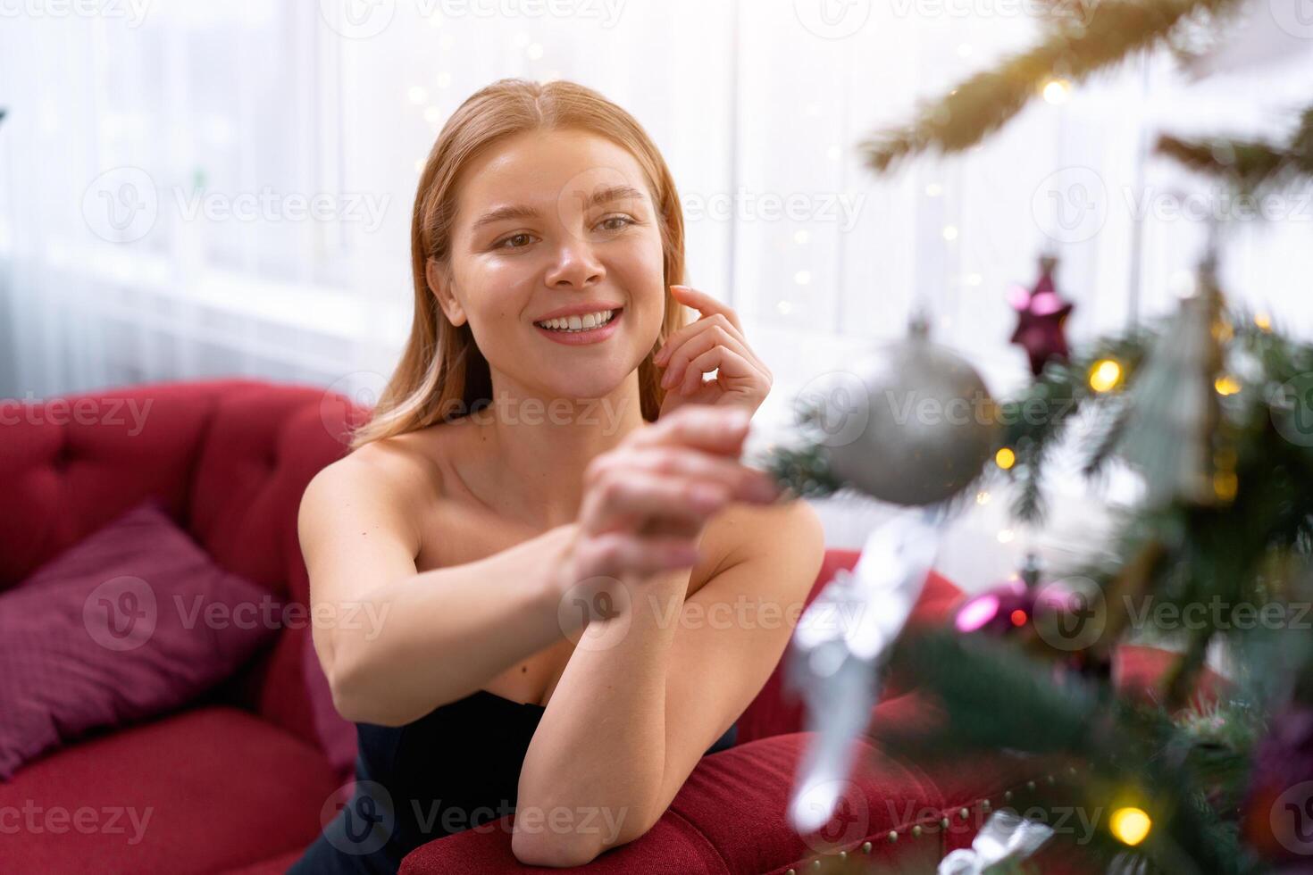 Young beautiful woman sitting sofa near Christmas tree looking on Christmas ornament and smile dressed black elegant sensitive dress with naked shoulders with big window background photo