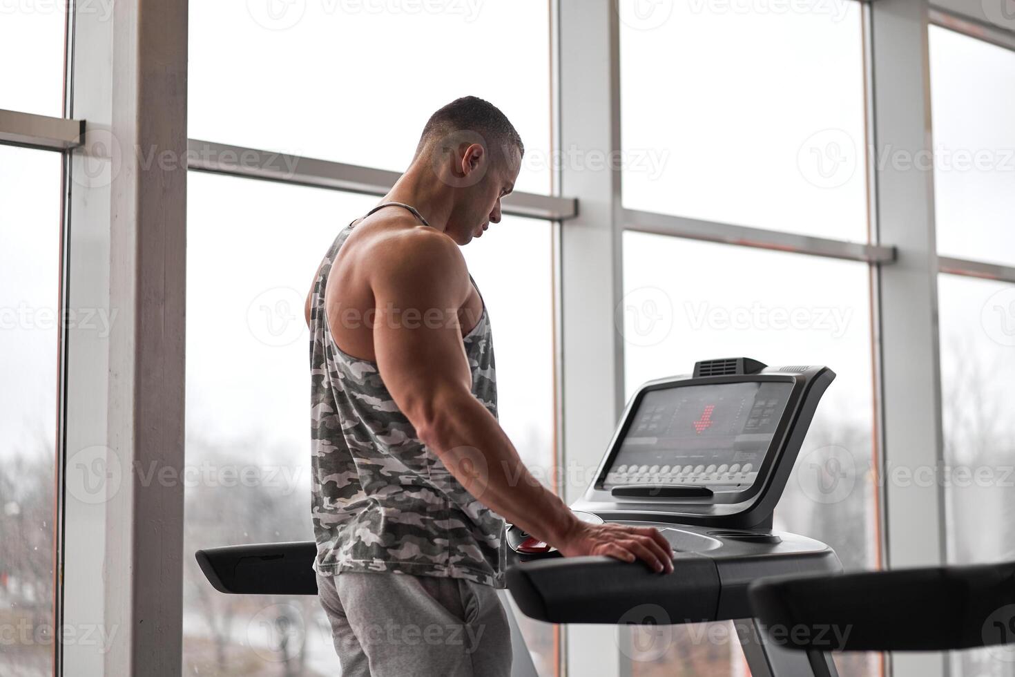 Muscular athletic bodybuilder fitness model running treadmill gym near big window photo