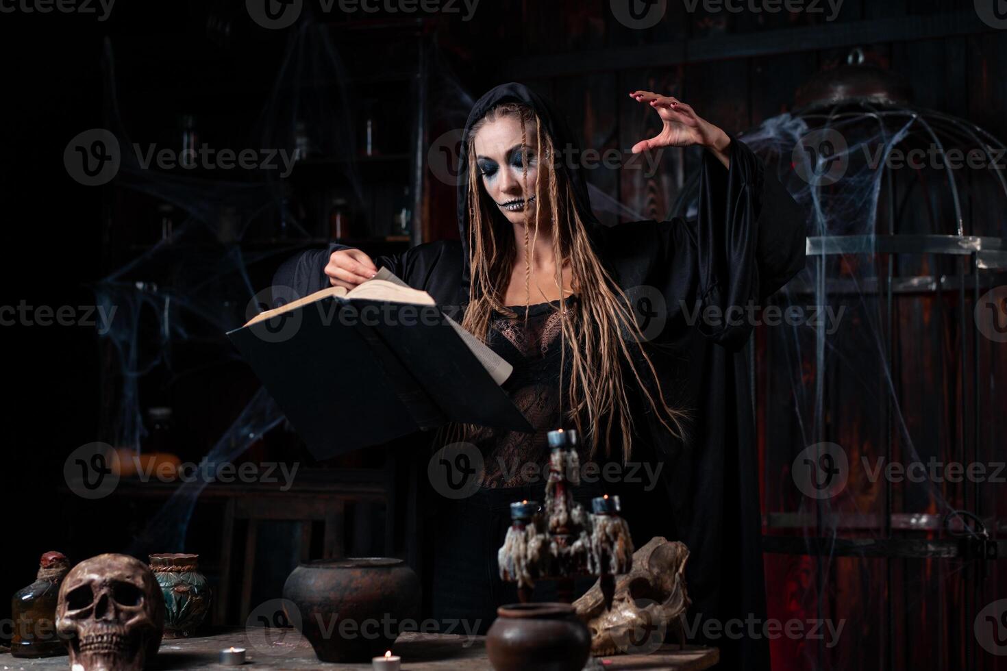 Halloween concept. Witch dressed black hood with dreadlocks standing dark dungeon room use magic book for conjuring magic spell photo