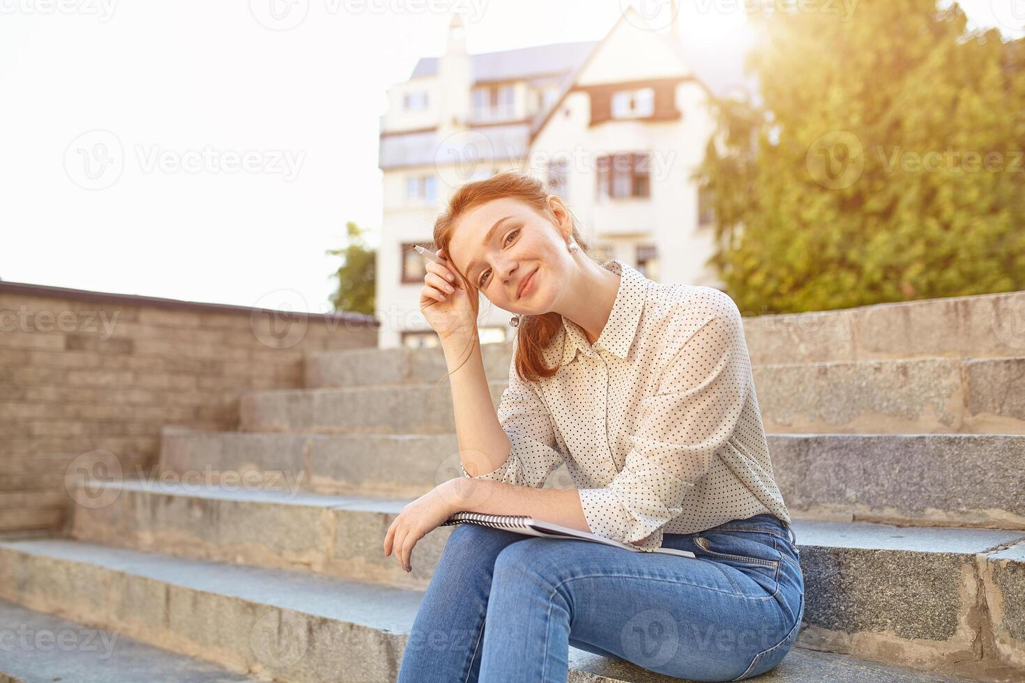 young beautiful student writes an essay in her notebook sitting on the steps stairs outdoor Red-haired girl with freckles photo