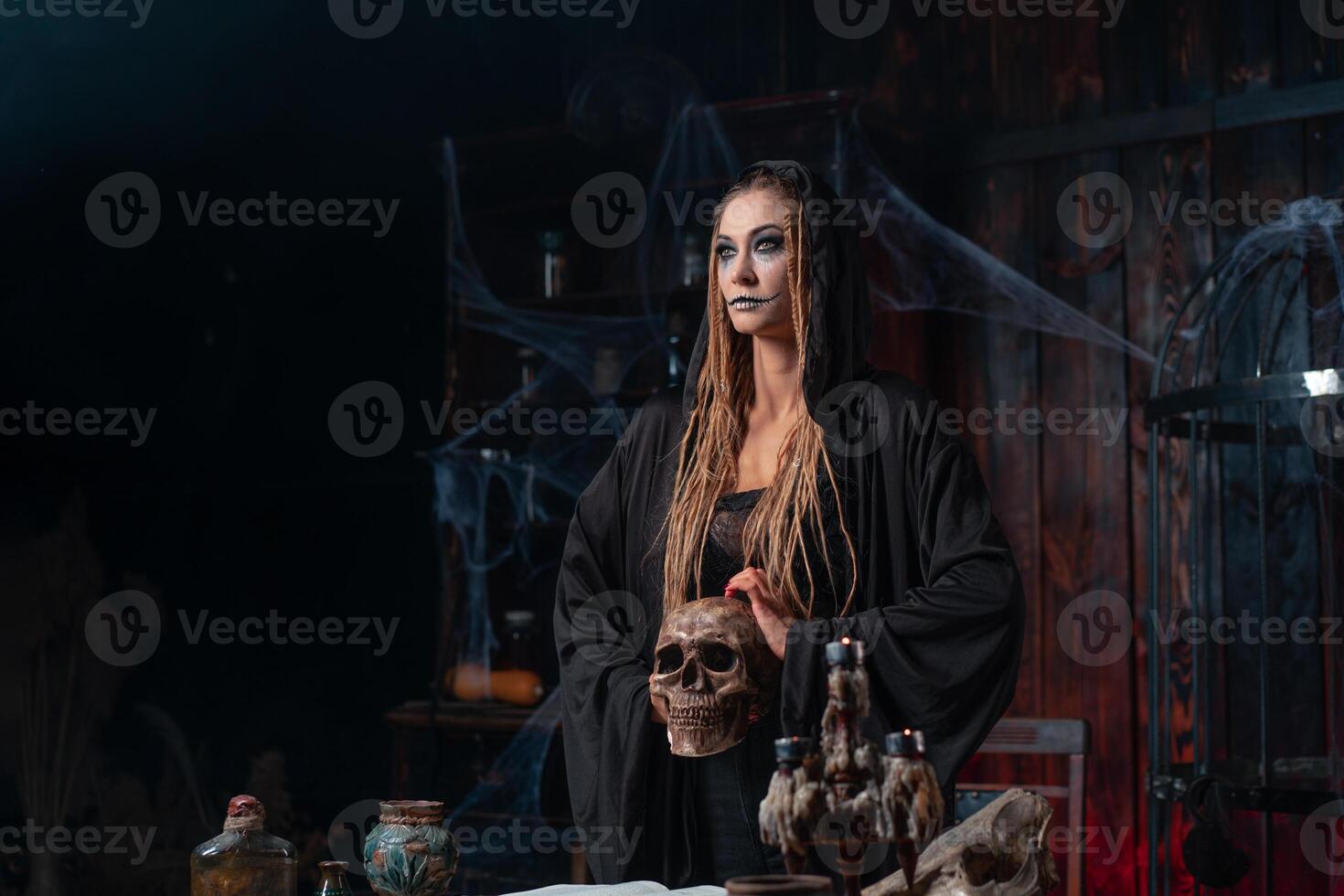 Halloween concept. Witch portrait close up with dreadlocks dressed black hood standing dark room with cage on background photo