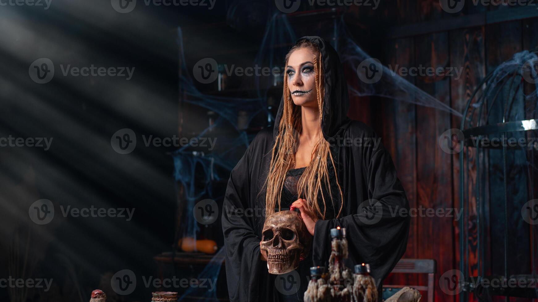 Halloween concept. Witch portrait close up with dreadlocks looking away dressed black hood standing dark room with cage on background photo