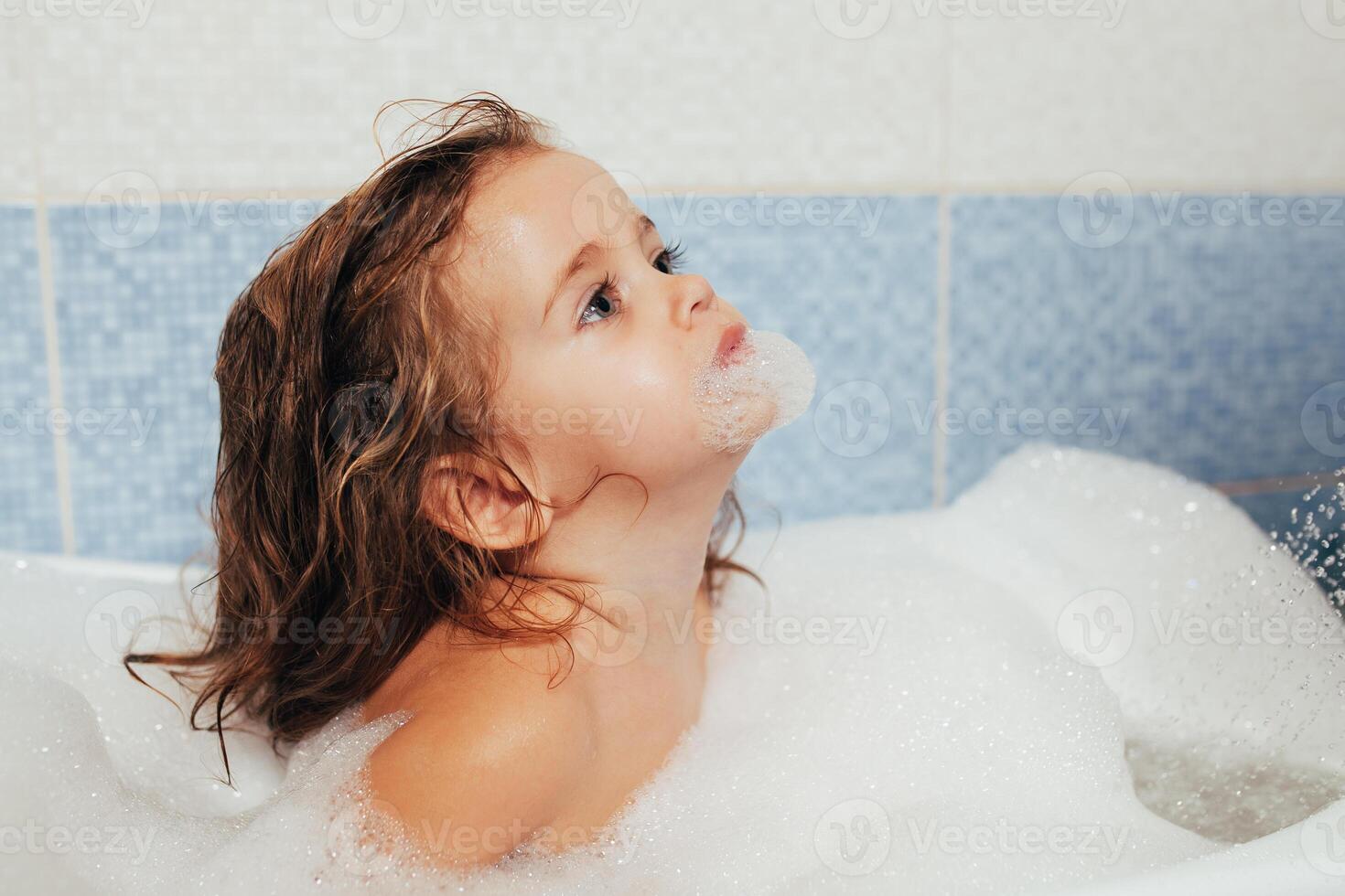 Fun cheerful happy toddler baby taking a bath playing with foam bubbles. Little child in a bathtub. Smiling kid in bathroom on blue background. Hygiene and health care. photo