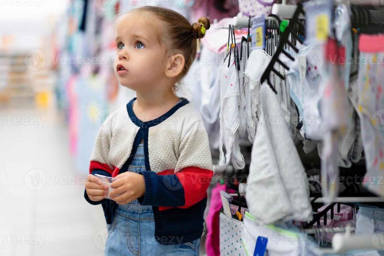 A little Caucasian girl is standing near a shop window with clothes and underwear in a large store. Little buyer. photo