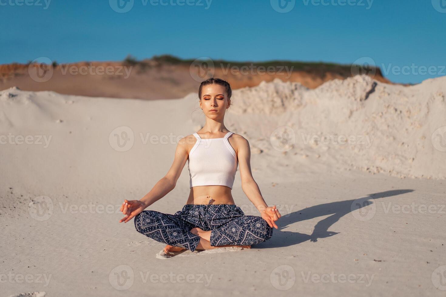 A beautiful young Caucasian girl in a white top and wide pants sits in a lotus position on the beach on the sand. photo