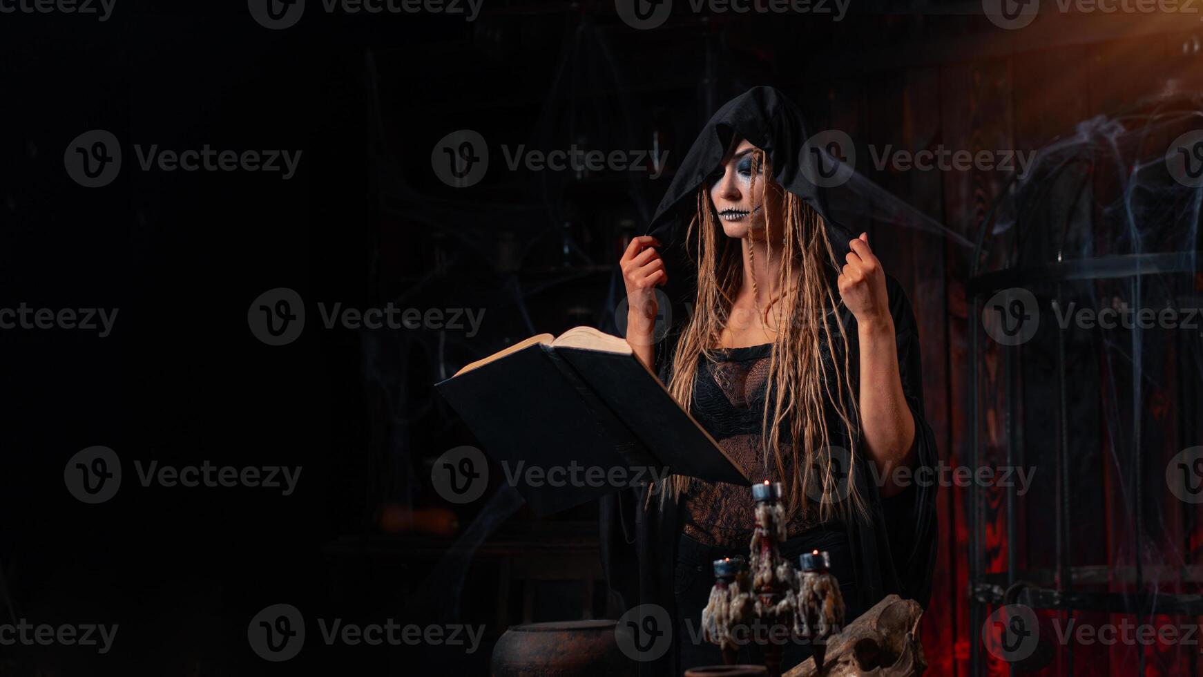 Víspera de Todos los Santos concepto. bruja vestido negro capucha con rastas en pie oscuro calabozo habitación utilizar magia libro para prestidigitación magia deletrear foto