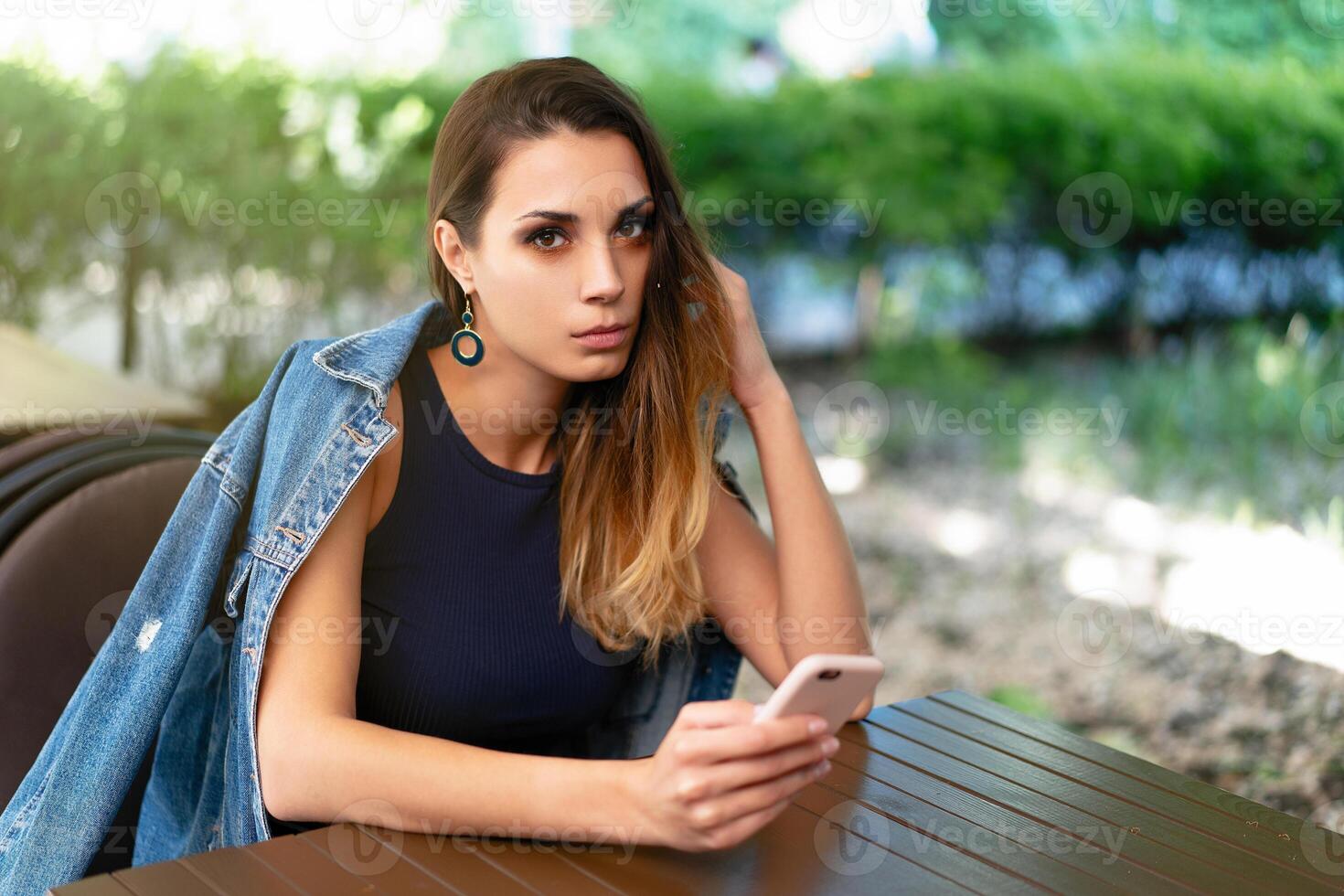 A lonely sad Caucasian girl sits in an open summer cafe with a phone in her hands and is sad alone photo