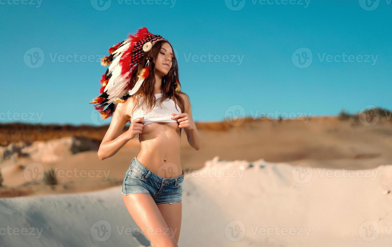 A beautiful young Caucasian girl in a white top and denim shorts on her head is wearing an Indian hat. Roach is in the desert. Happy holiday mood. photo