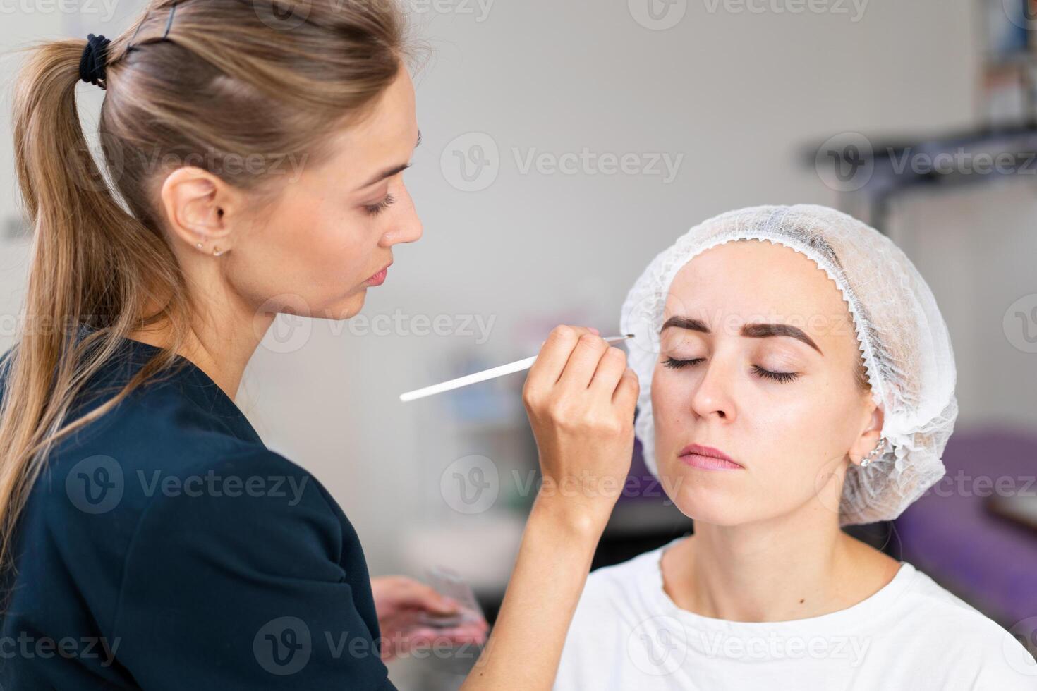 Cosmetology cabinet client sitting on couch. Beautician applies marking on eyebrows. photo