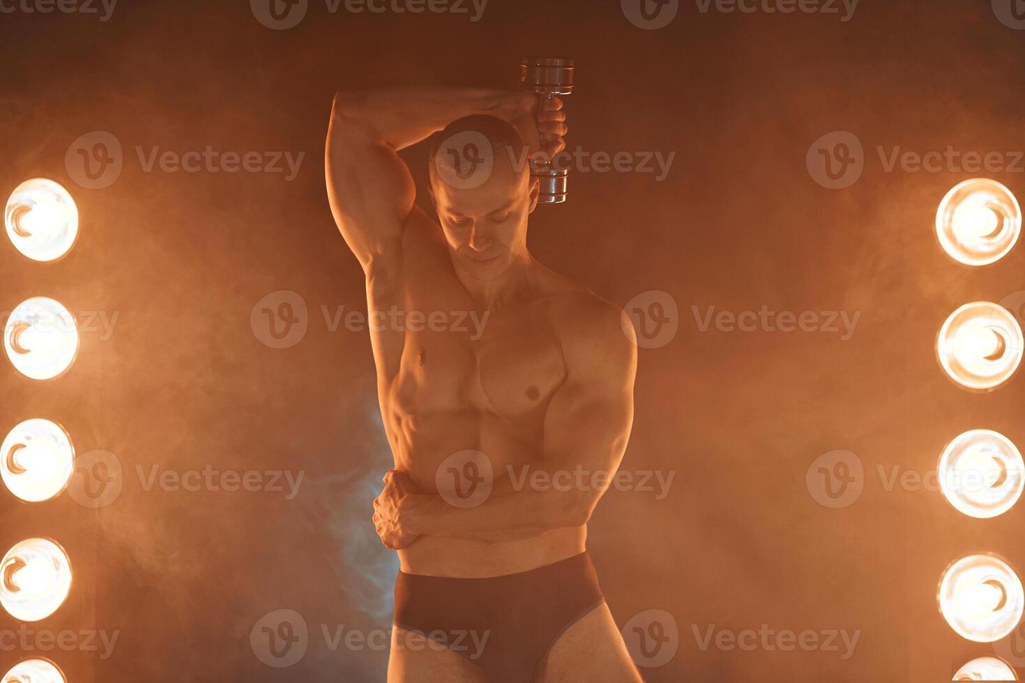 Young pumping dumbbells, practicing triceps training, smoky illuminated background photo