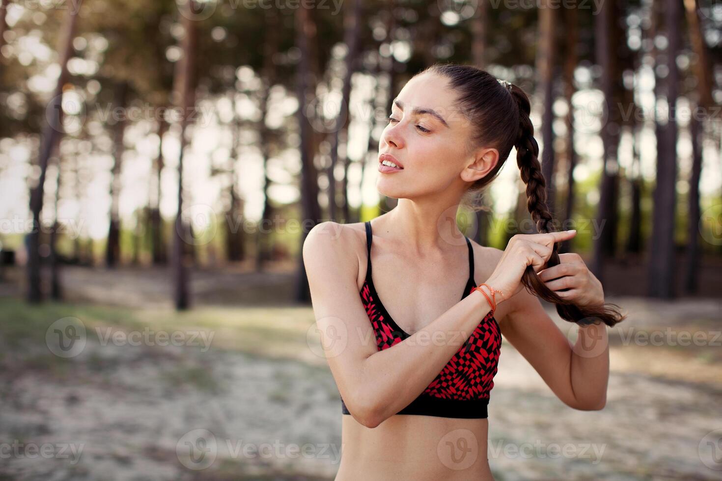 People, sports, nature and fitness. Close up portrait of young woman runner with long braid dressed in sportswear standing on trail in green forest photo