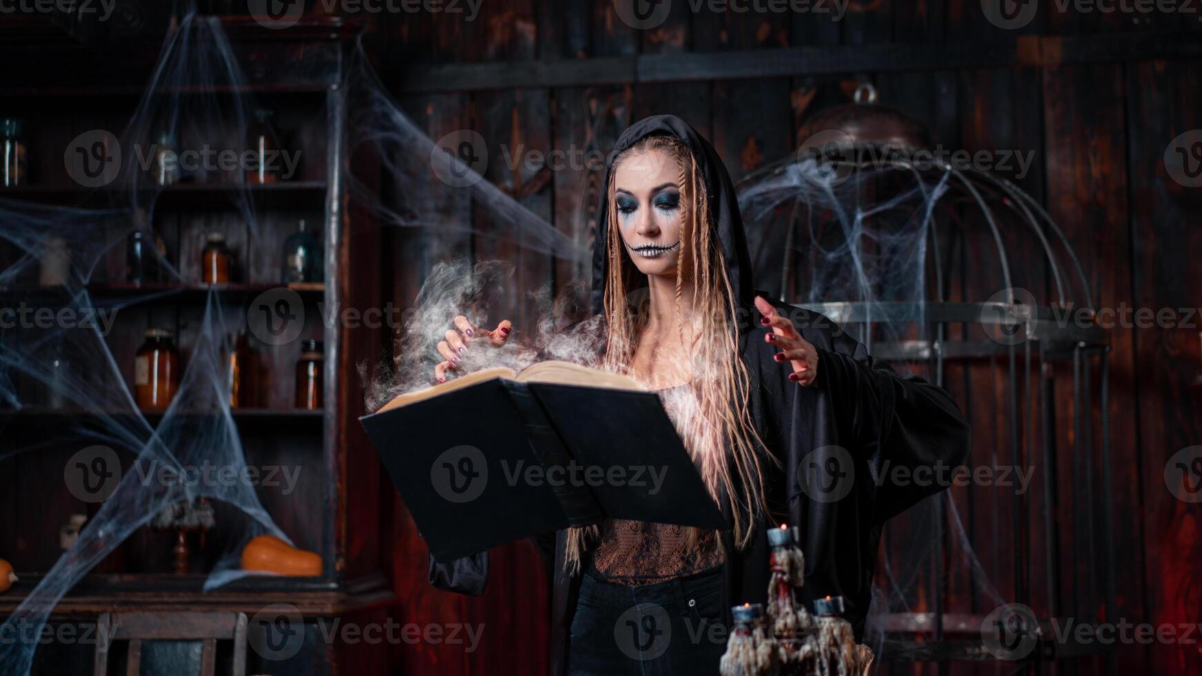 Víspera de Todos los Santos concepto. bruja vestido negro capucha con rastas en pie oscuro calabozo habitación utilizar magia libro para prestidigitación magia deletrear foto