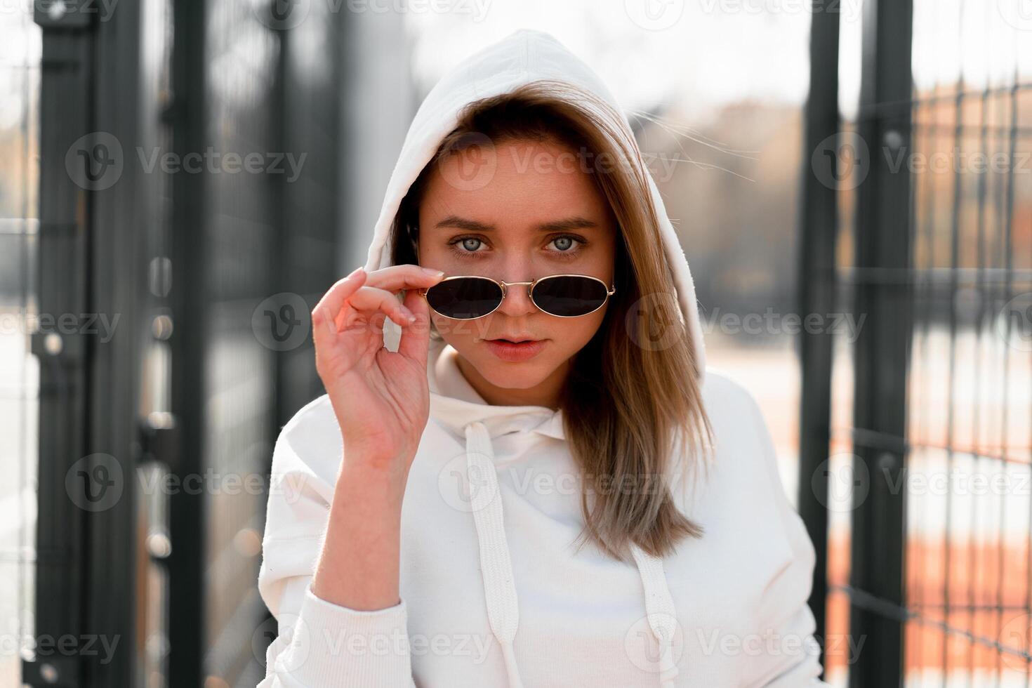 Outdoor close up portrait of young beautiful woman with long hair in sunglasses, dressed in a white sweater, near the sportsground photo