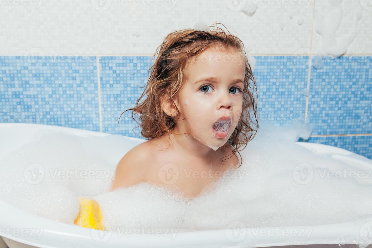 Fun cheerful happy toddler baby taking a bath playing with foam bubbles. Little child in a bathtub. Smiling kid in bathroom on blue background. Hygiene and health care. photo