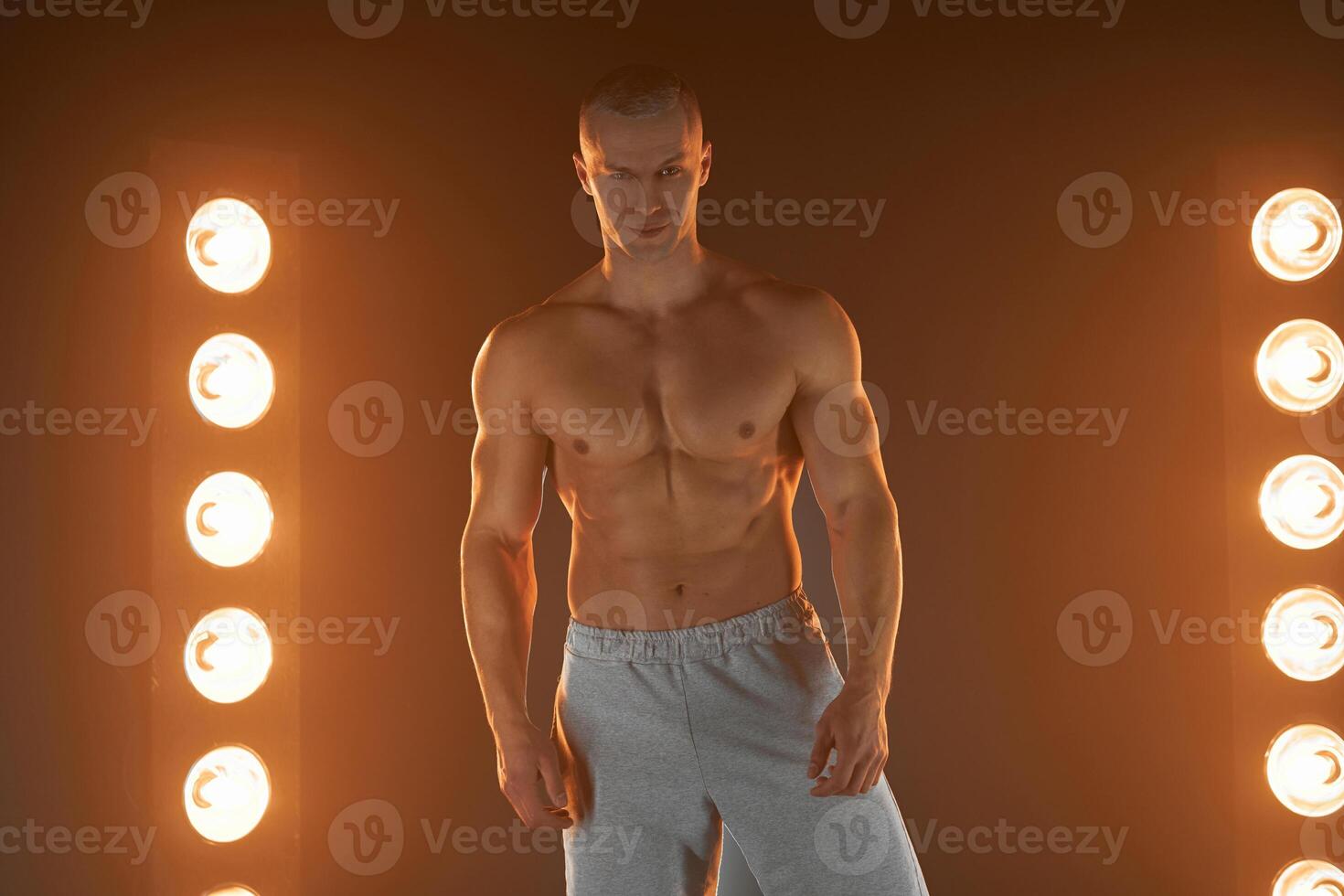 Active lifestyle. Professional bodybuilder showing perfect muscular body and smiling at camera, lamps illumination on background photo