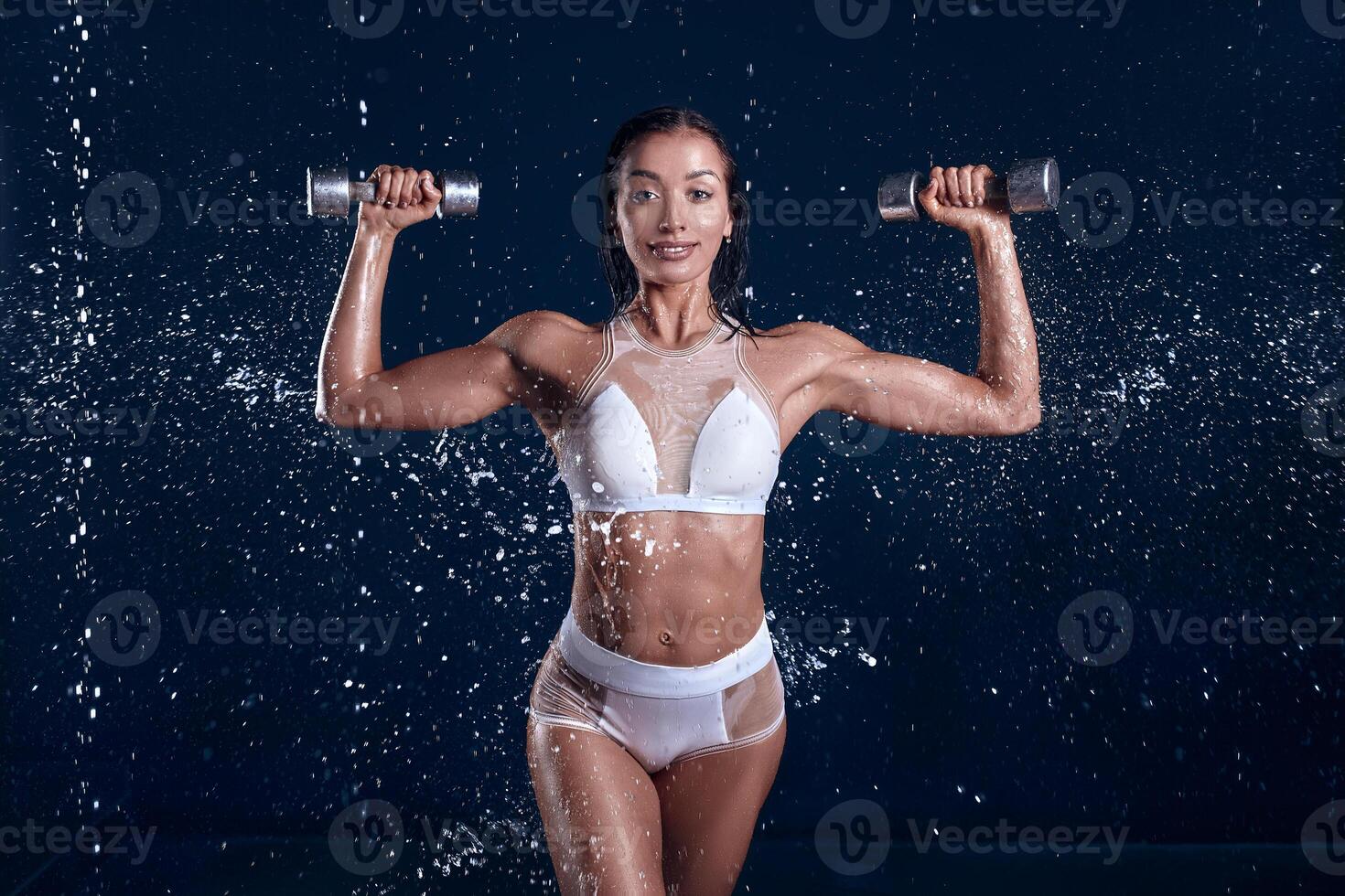 Beautiful young girl in sportswear poses with dumbbells in aqua studio. Drops of water spread about her fitness body. The perfect figure on the background of water splashes photo