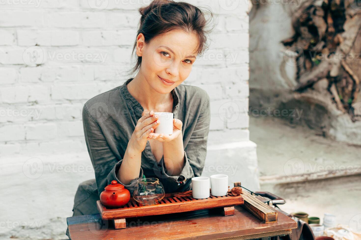 Oriental Master of tea ceremony with white brick wall on the background. Traditional tea party on the nature with woman dressed in the gray shirt photo