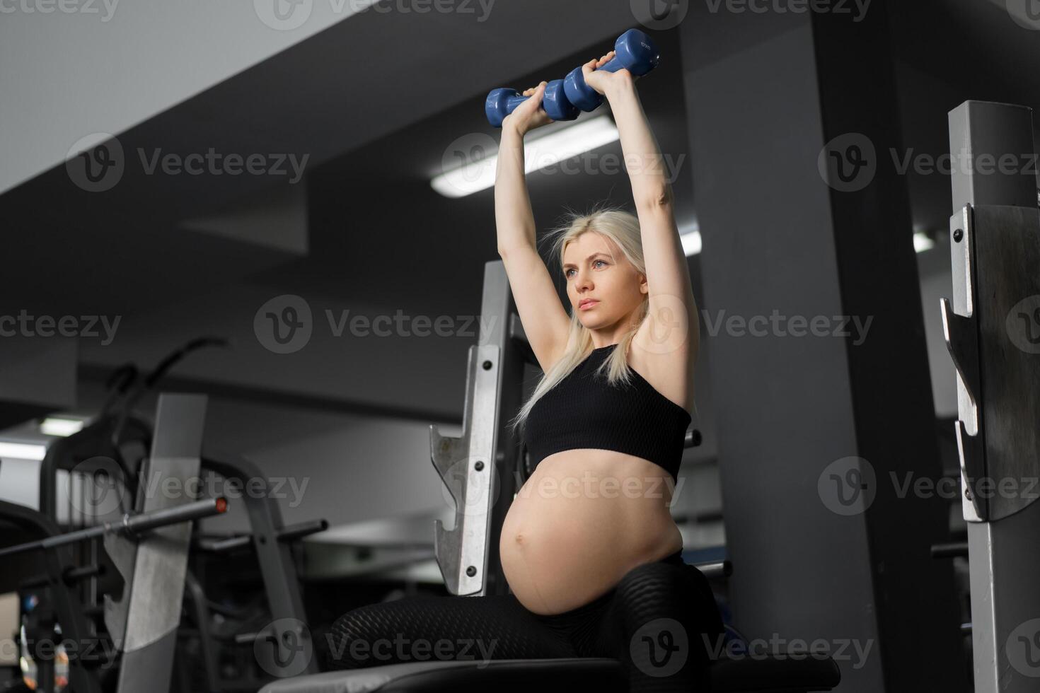 Pregnant woman lifting up dumbbells training arm and shoulders muscle at the gym sitting bench photo