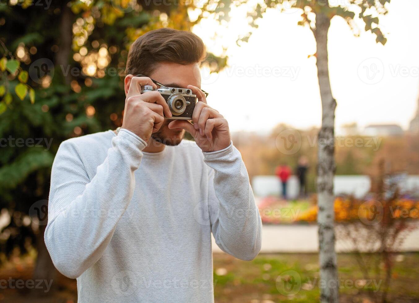 Young handsome hipster guy walks in a beautiful autumn park on the background of yellow leaves in warm sunny weather and takes pictures on a pig film camera. Autumn leisure time. Creative youth photo