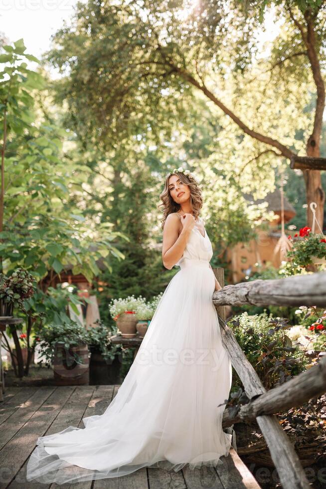 A cute curly woman in a white wedding dress with a wedding bouquet and wreath in her hair standing back to the camera in nature. Concept escaped bride. Forward to a happy bright future Runaway photo