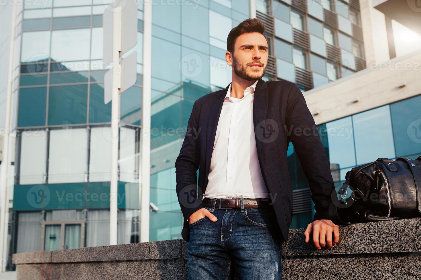handsome young businessman with a beard and in a business suit standing on the street against the background of the office building next to a comfortable stylish leather bag. photo
