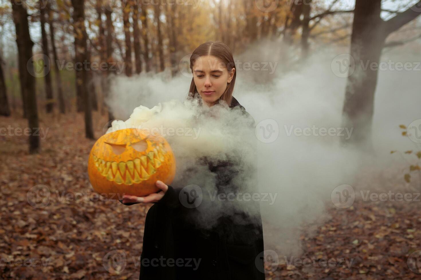 un hermosa niña con largo pelo en un negro capa es participación un naranja calabaza desde cuales fumar es próximo. contento Víspera de Todos los Santos. oscuro otoño bosque. horrores foto