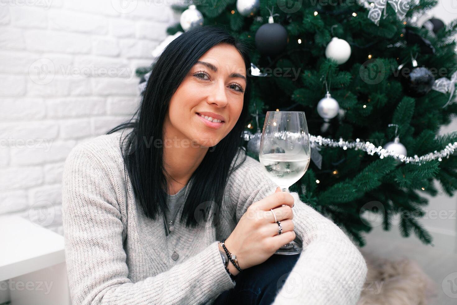 A young woman sits near a Christmas tree, holding a glass and a Christmas toy. Preparation for the New Year's celebration photo