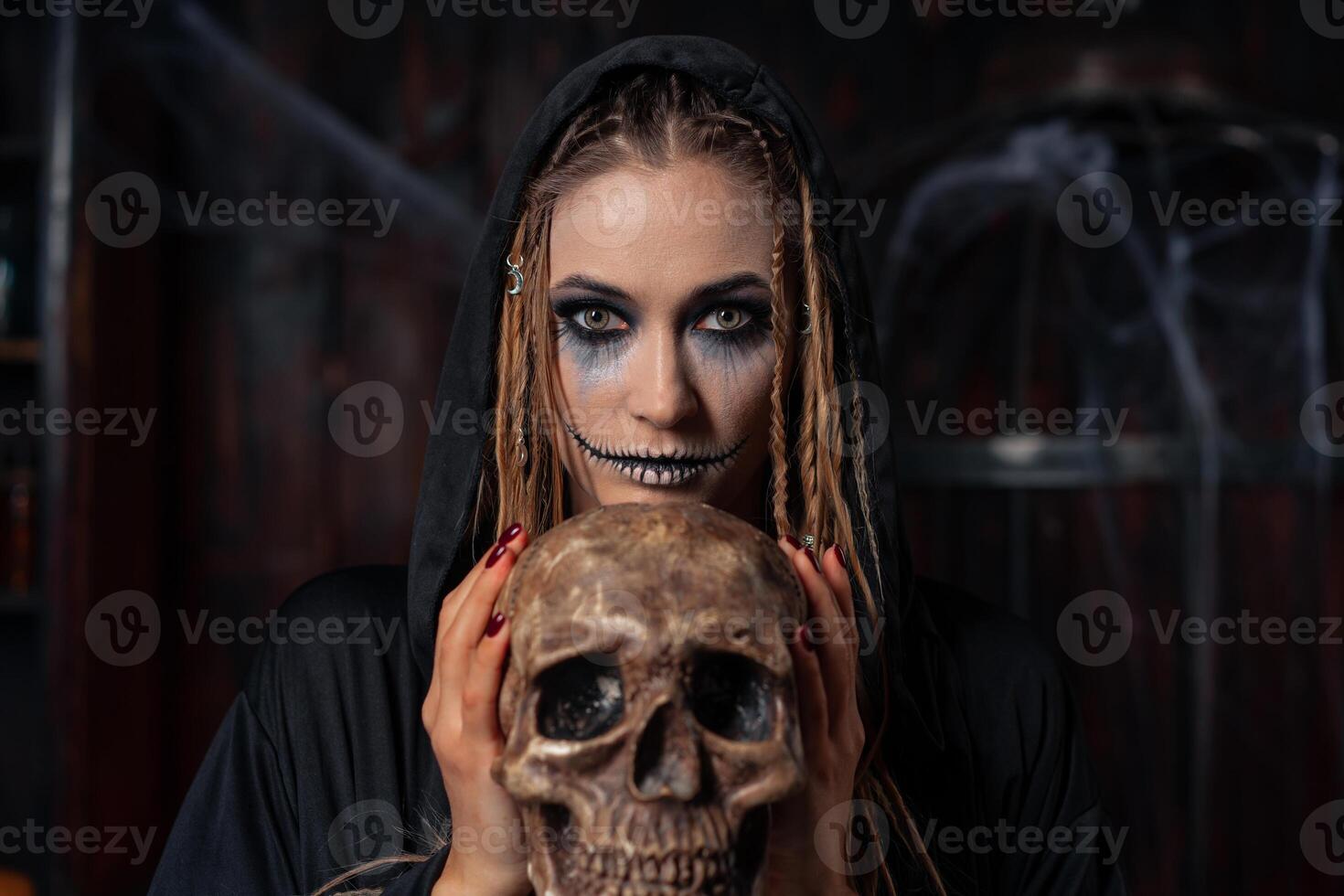 Víspera de Todos los Santos concepto. bruja retrato cerca arriba con rastas mirando cámara vestido negro capucha en pie oscuro habitación con jaula en antecedentes foto