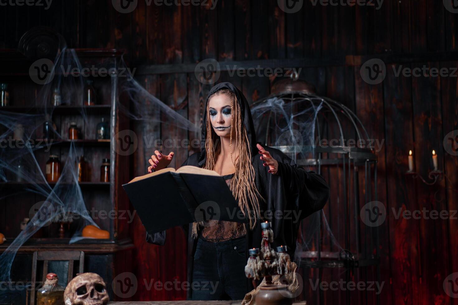 Halloween concept. Witch dressed black hood with dreadlocks standing dark dungeon room use magic book for conjuring magic spell photo