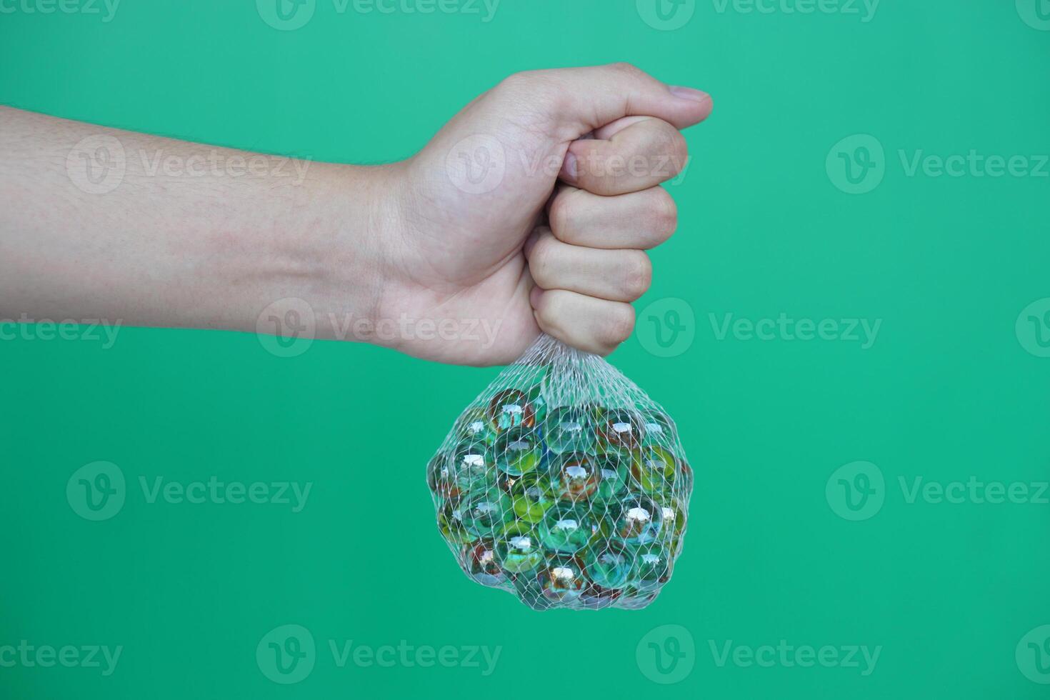 Close up hand hold net bag of marbles balls, small spherical object often made from glass. Green background. Concept. object use as toy for playing games or use for DIY craft decoration. photo