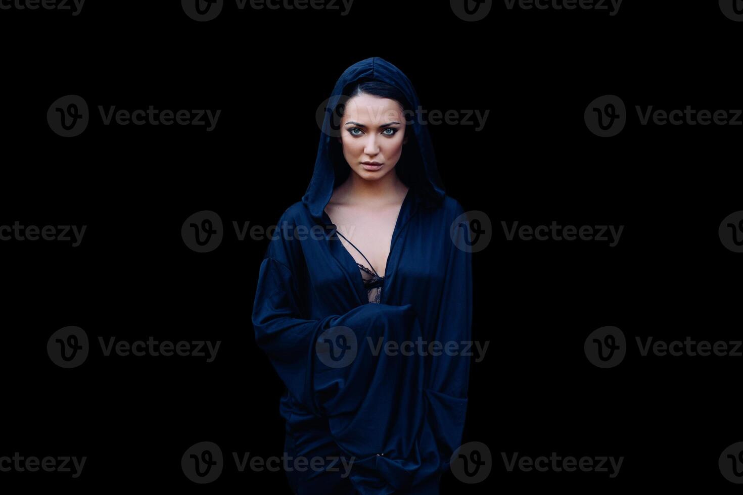 Young beautiful woman with a black hair and in the dark blue cloak with hood at the black background photo