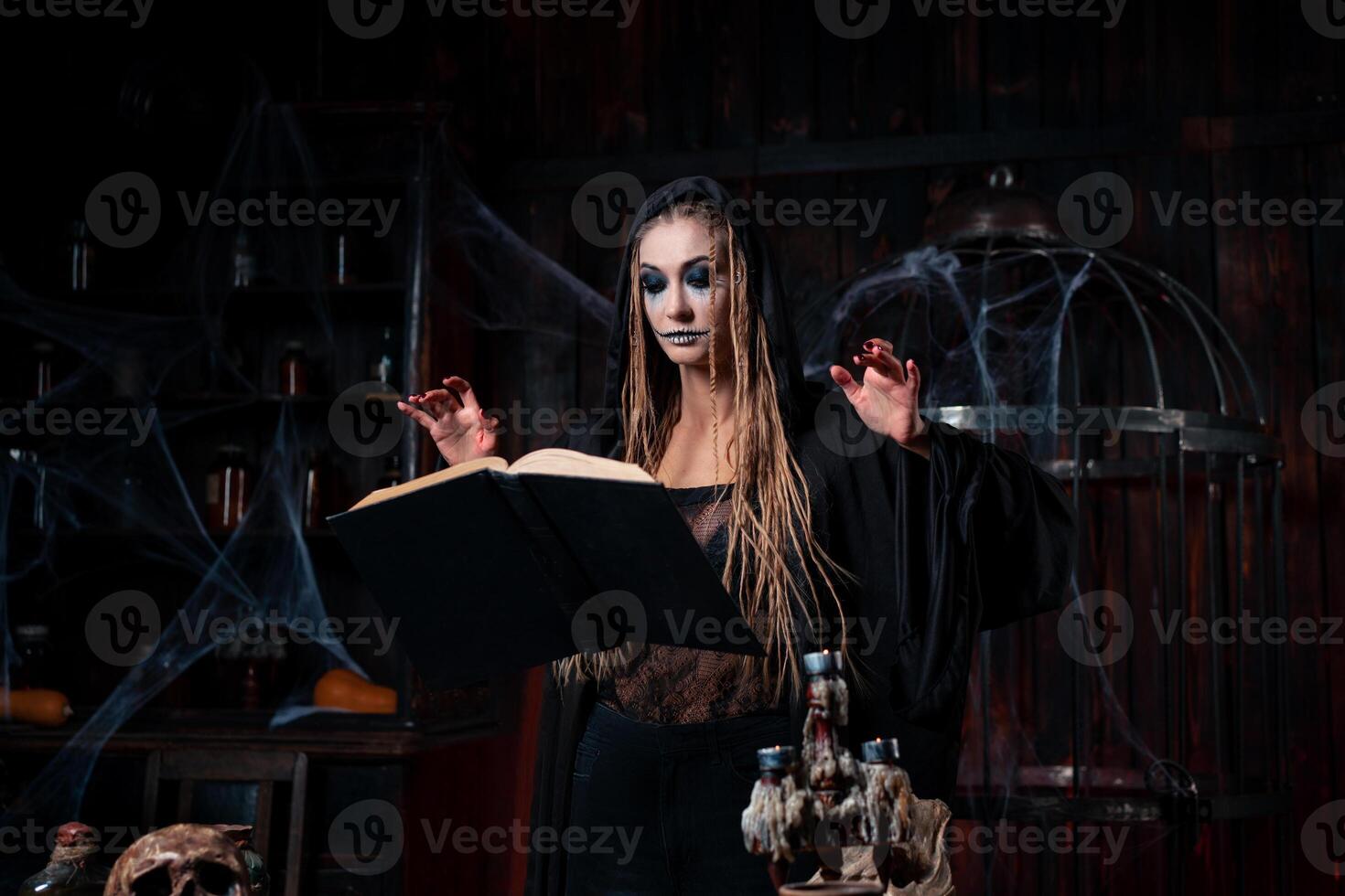 Halloween concept. Witch dressed black hood with dreadlocks standing dark dungeon room use magic book for conjuring magic spell photo