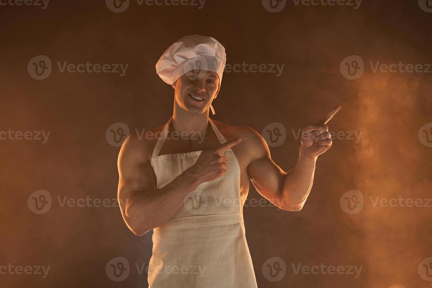 New normal concept. Muscular chef wearing protective medical mask apron and chef hat, standing show finger side on smoky background and lamp illumination photo