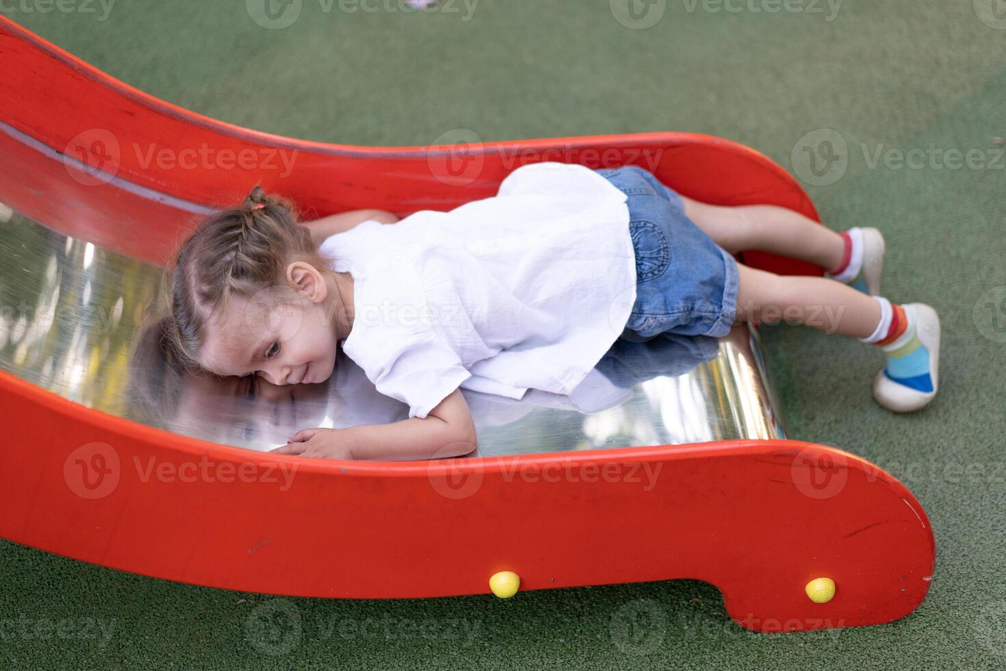 pequeño caucásico niña mentiras en un diapositiva a el patio de recreo cansado de un triste verano día. foto