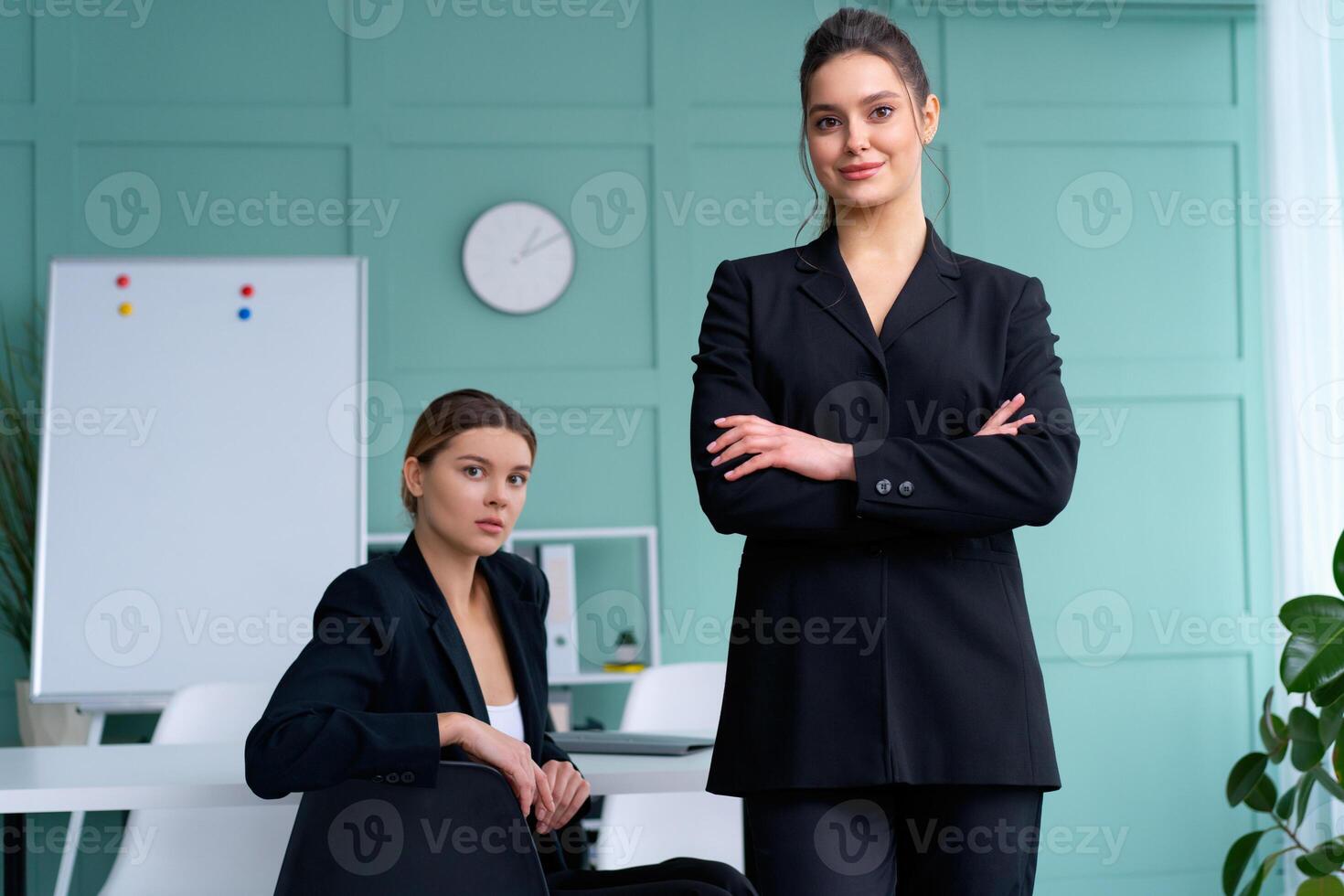 Two young women leaders dressed black suit in office one woman standing arms crossed other woman sitting on the table looking at camera. Business meeting photo