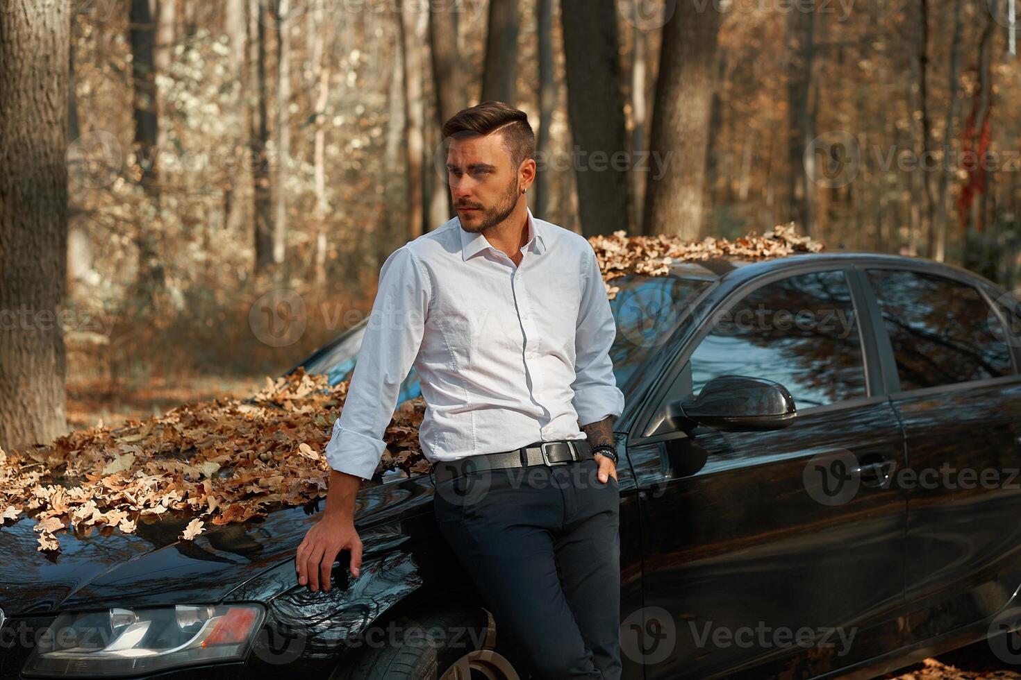 un hermoso joven caucásico hombre vistiendo negocio traje en pie cerca negro coche en naturaleza. foto