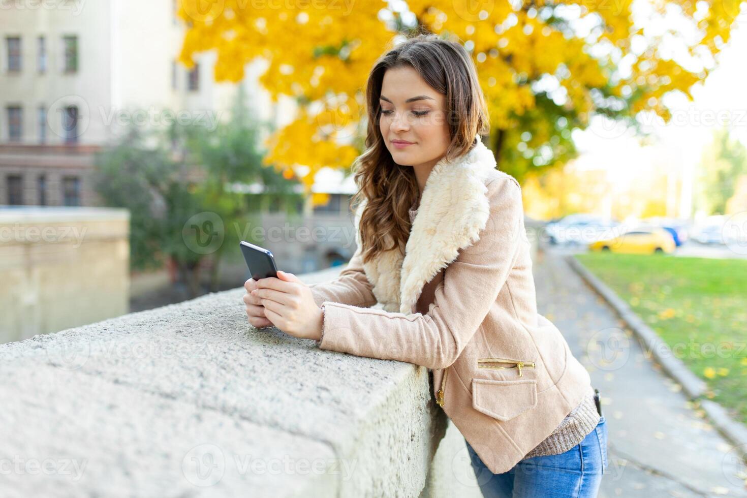 Beautiful caucasian brunette girl standing warm autumn day with background of trees with yellow foliage and a city. Dressed wool sweater and jacket. photo