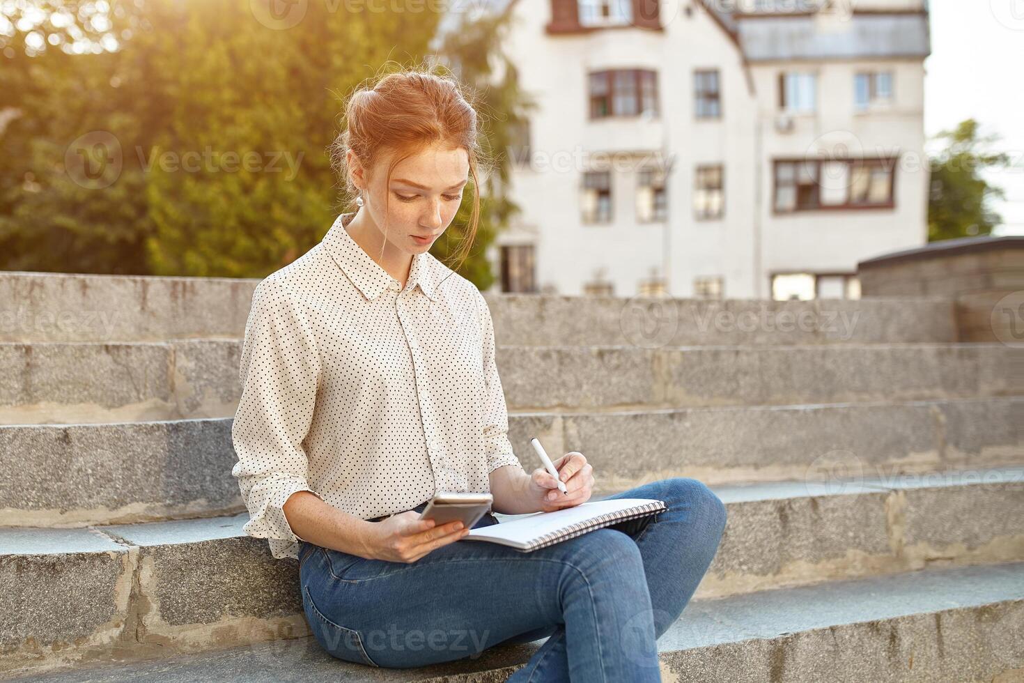 young beautiful student writes an essay in her notebook sitting on the steps stairs outdoor Red-haired girl with freckles photo