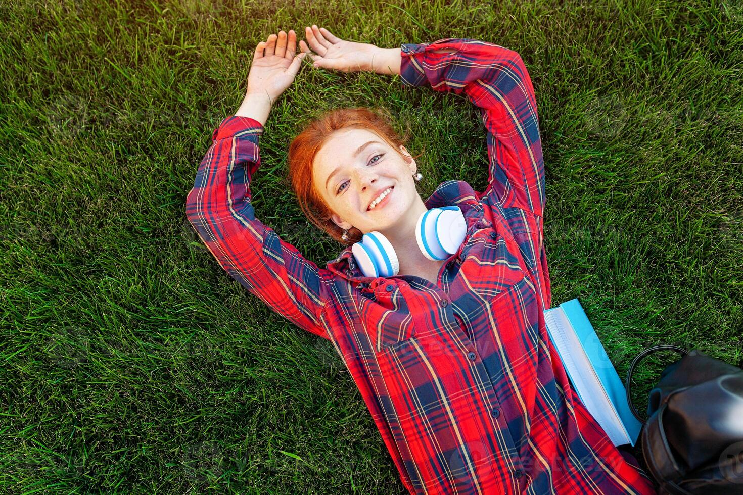 Beautiful young red-haired student girl with freckles lying on her back on grass and lawn view from above. Dressed in a red checkered shirt, resting after classes photo