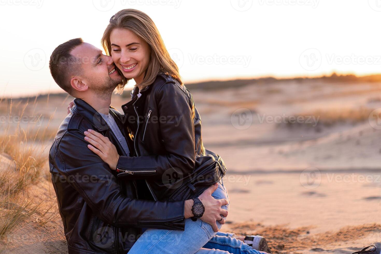 Beautiful young Caucasian couple dressed black leather jacket and blue jeans. A man sits on the sand woman sits on top and hugs him. photo