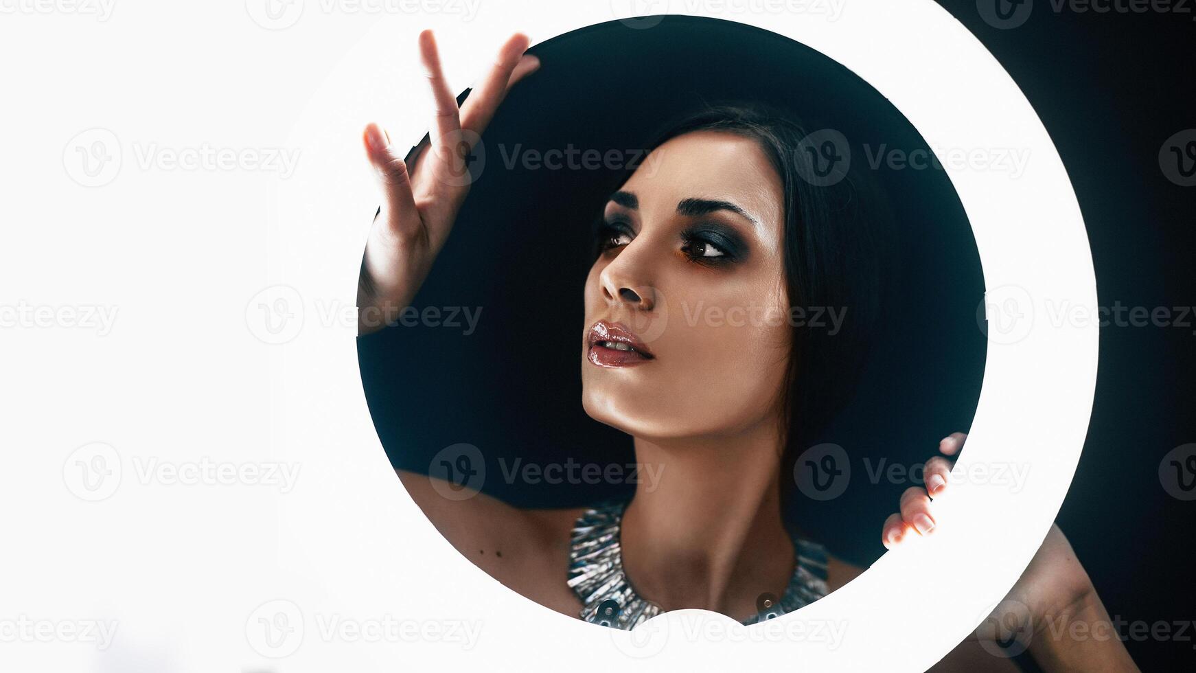 A beautiful young Caucasian girl with a beautiful make-up looks out from behind a circular lamp for portrait photo shooting