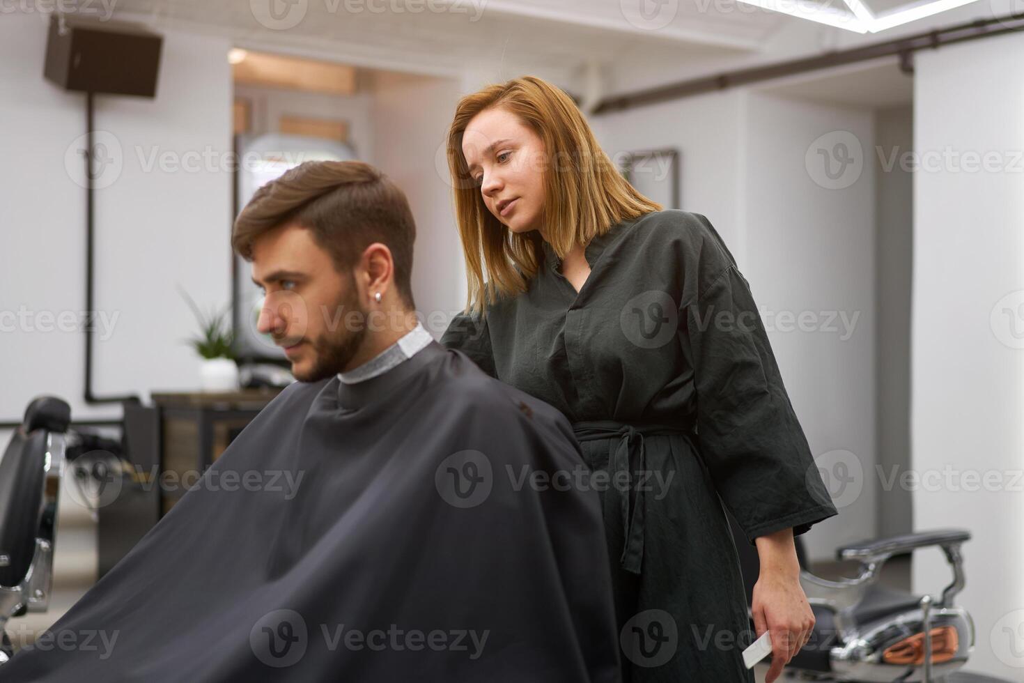 Handsome blue eyed man sitting in barber shop. Hairstylist Hairdresser Woman cutting his hair. Female barber. photo