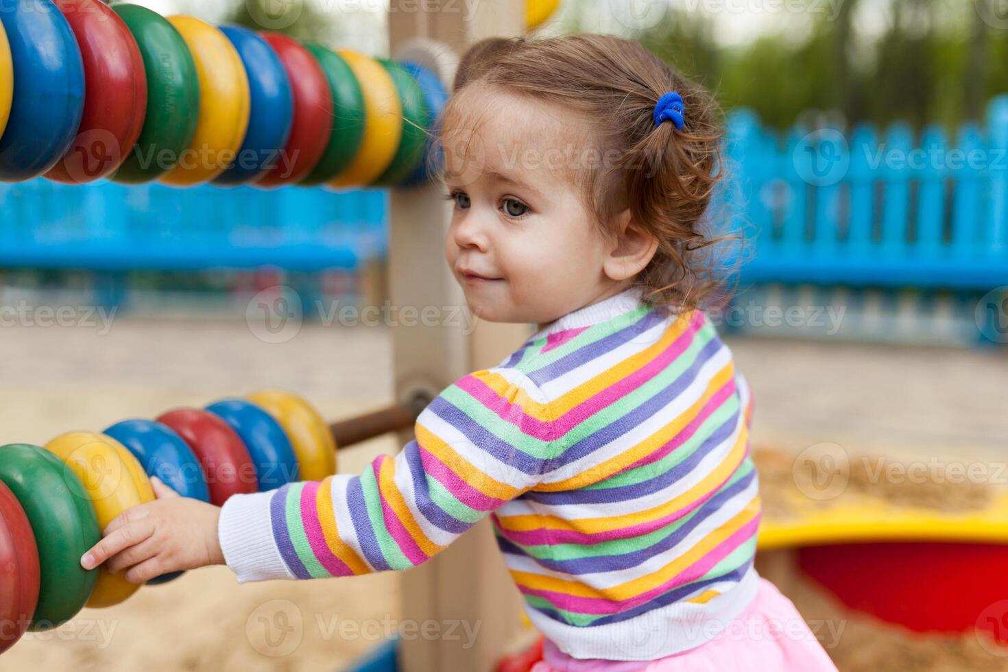 a little girl with two tails is dressed in a striped colorful jacket is playing in the sandbox on the playground photo