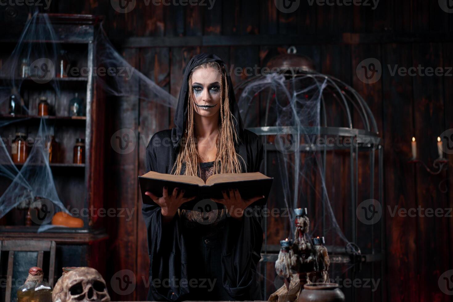 Halloween concept. Witch dressed black hood with dreadlocks standing dark dungeon room use magic book for conjuring magic spell photo