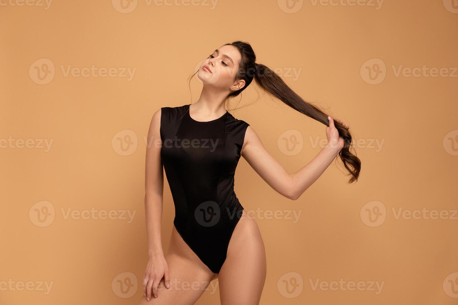Young beautiful woman dressed seductive black bodysuit standing studio isolated on color background holding hair ponytail looking away photo