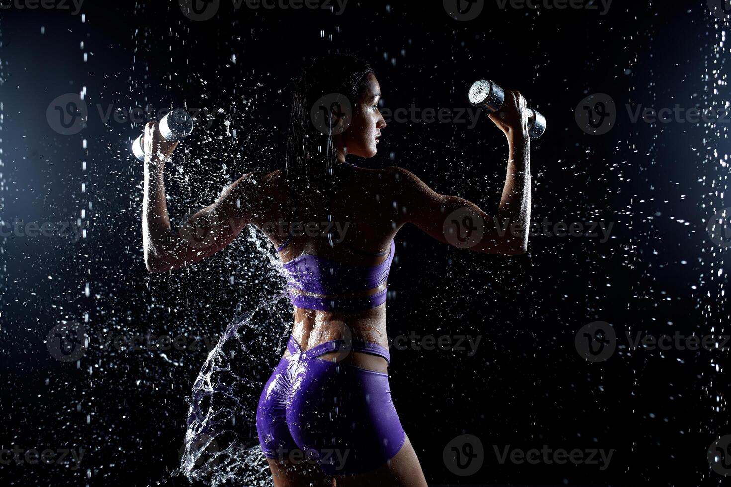 Beautiful young girl in purple sportswear poses with dumbbells in aqua studio. Drops of water spread about her fitness body. The perfect figure on the background of water splashes photo