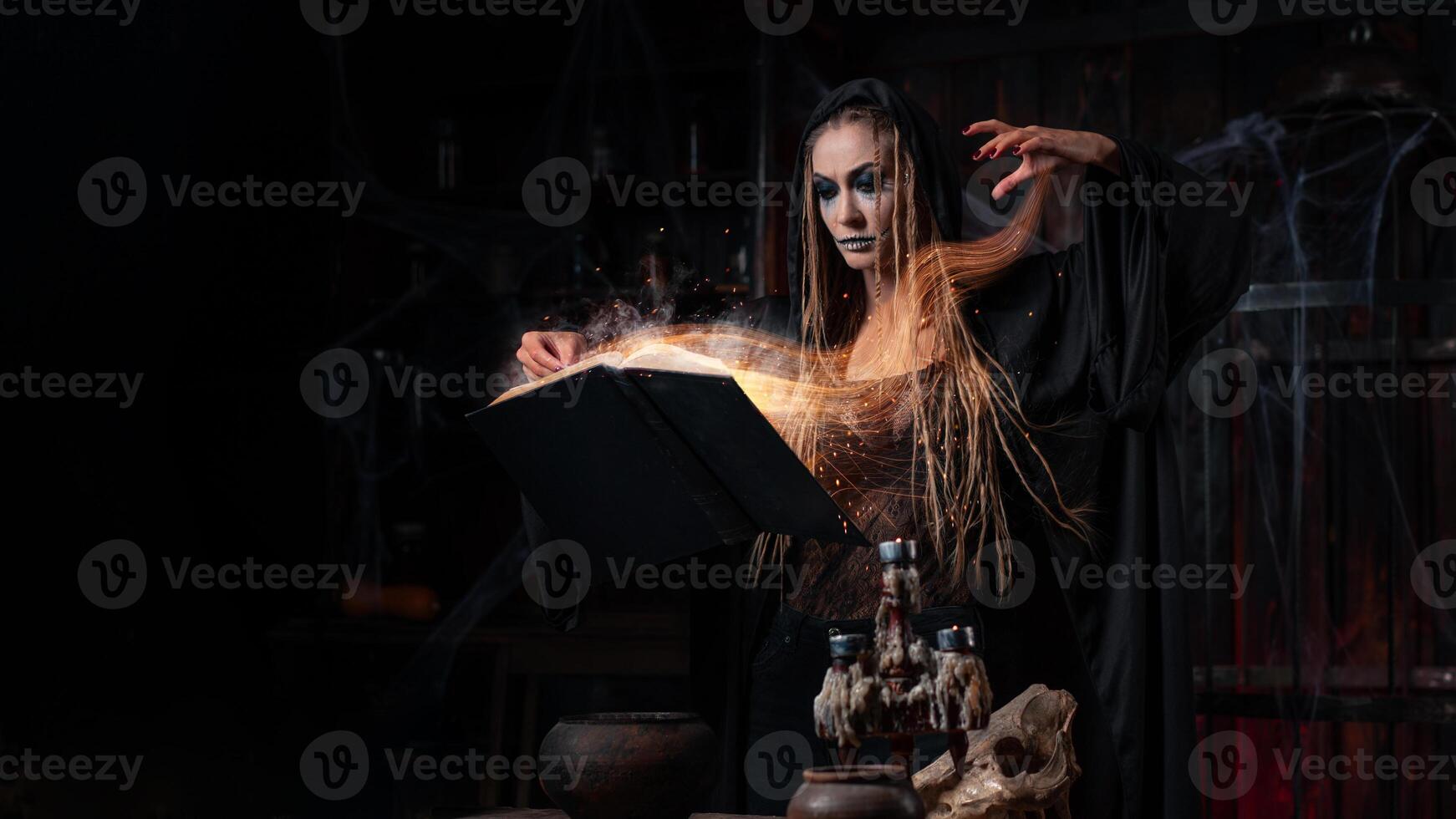 Halloween concept. Witch dressed black hood with dreadlocks standing dark dungeon room use magic book for conjuring magic spell photo