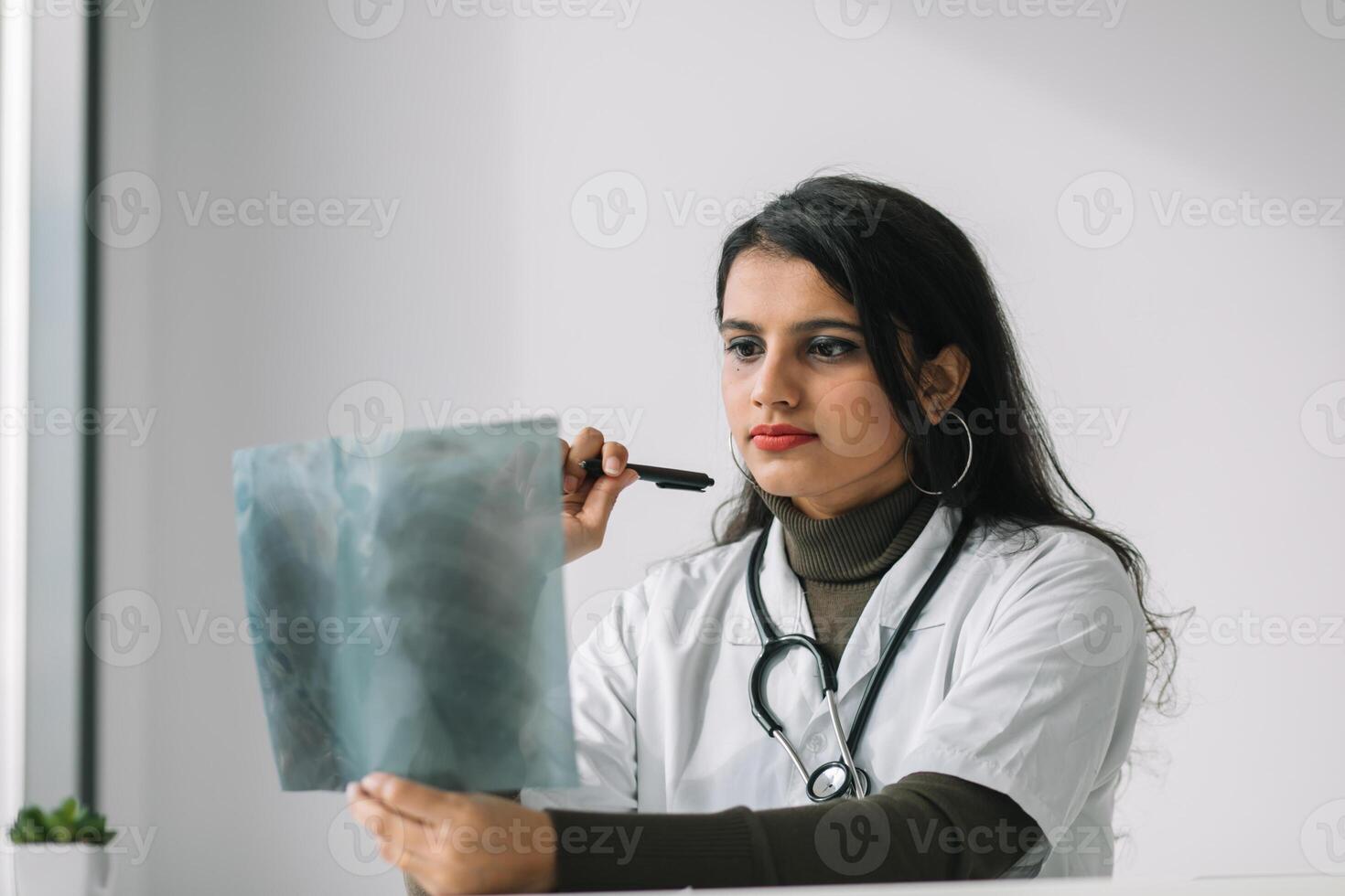 Indian female doctor in white coat with stethoscope examines x-ray and makes notes. Young indian medical student at the clinic in practice photo