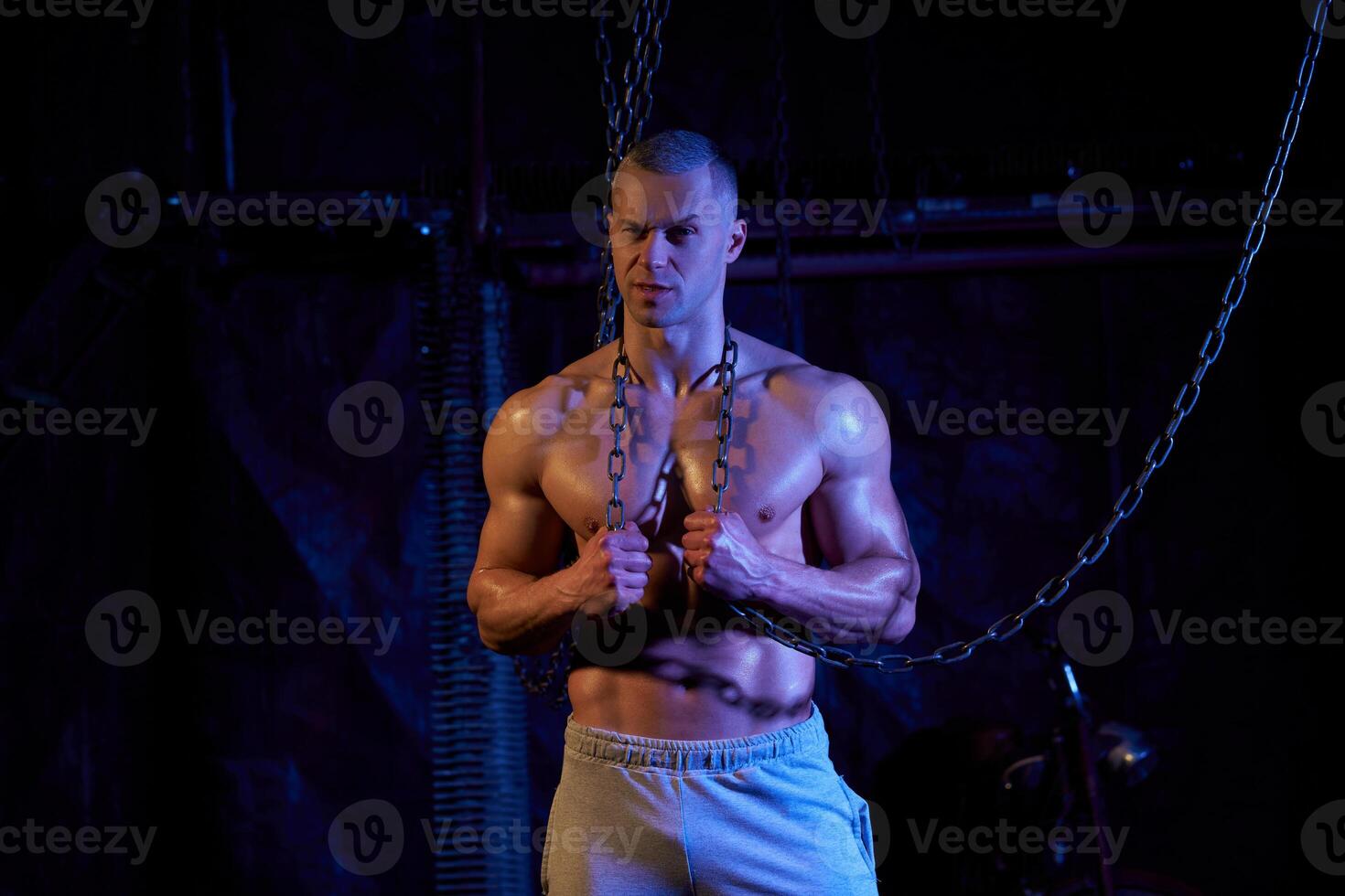 Young shirtless muscular man standing among metal chains, looking at camera, copy space photo