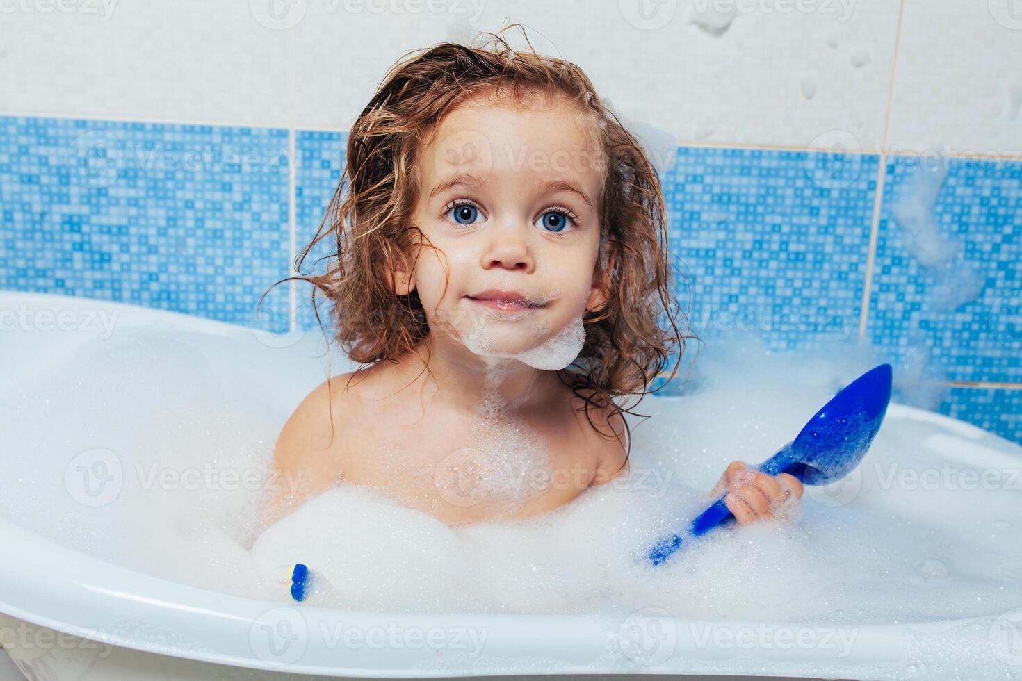 Fun cheerful happy toddler baby taking a bath playing with foam bubbles. Little child in a bathtub. Smiling kid in bathroom on blue background. Hygiene and health care. photo